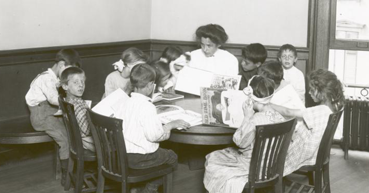In a photo from the turn of the century, a librarian and several children read books around a table