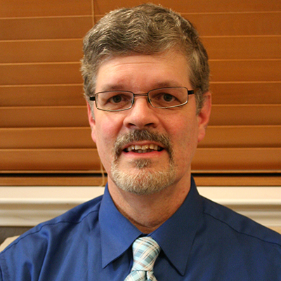 Photo of a man with gray hair and a mustache wearing a blue shirt and tie in front of a window