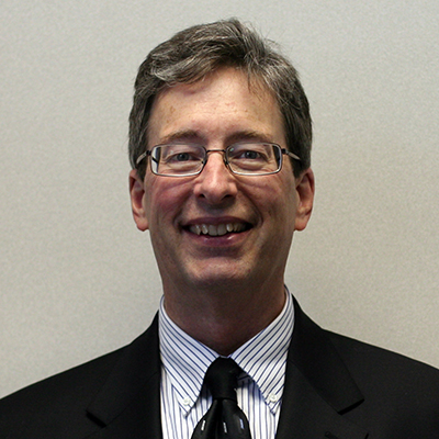 Photo of a man with gray hair and glasses wearing a suit and tie and smiling