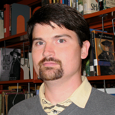 Photo of a man with dark hair and a goatee waring a gray sweater in front of library shelves