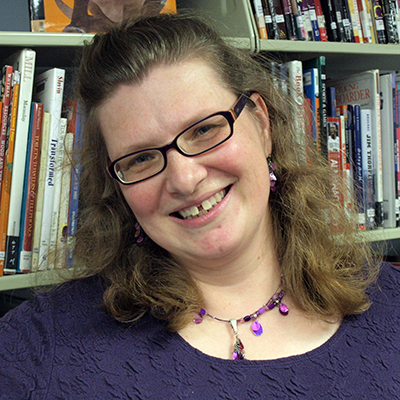 Photo of a woman with brown hair and glasses wearing a purple shirt and smiling in front of library shelves