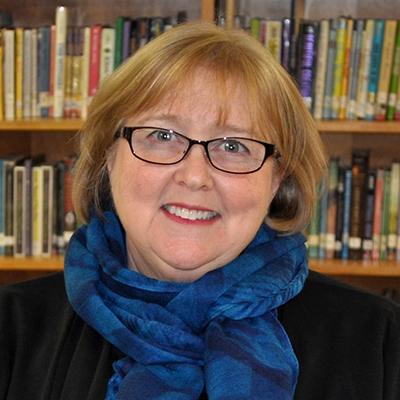 Photo of a woman with glasses and a blue scarf smiling in front of library shelves
