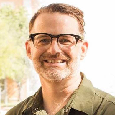 Photo of man with short hair and glasses in an army green shirt smiling outside