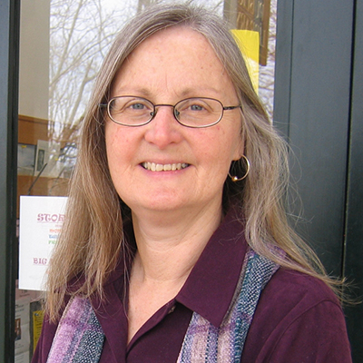 Photo of a woman with long gray hair and glasses wearing a purple shirt and scarf and smiling