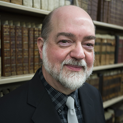 Photo of bald man with a gray beard and gray tie in front of library shelves