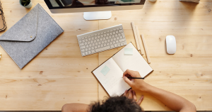 A person writing in a notebook in front of a desktop computer