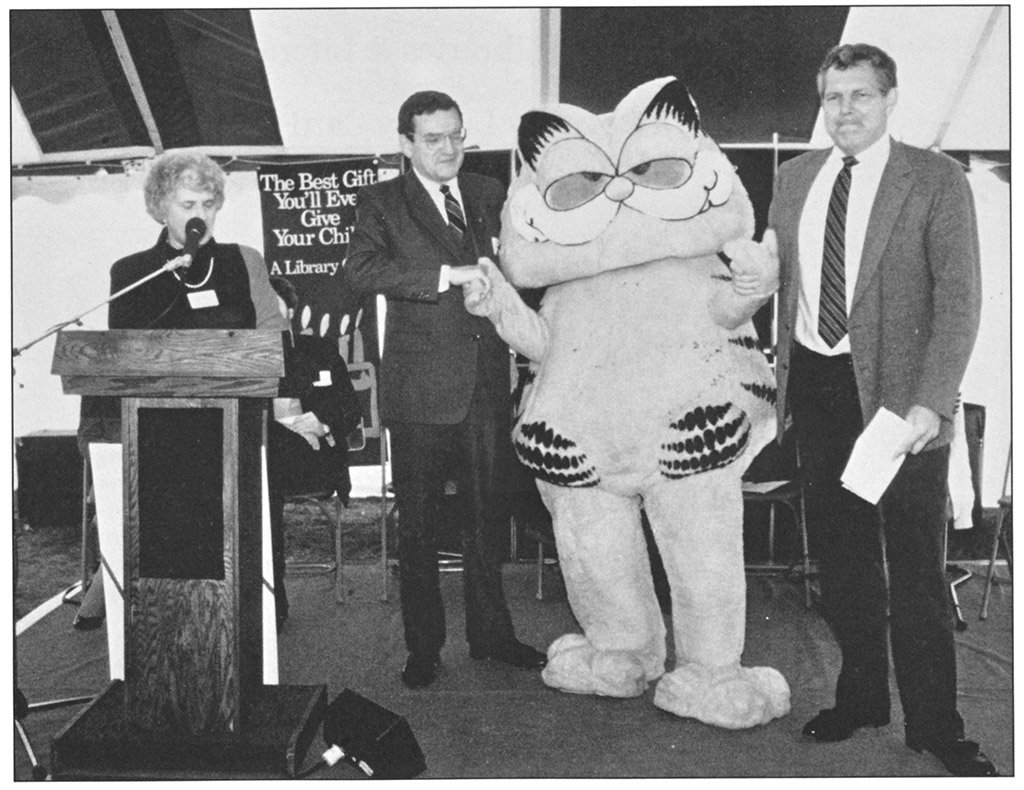 A black and white photo of several people standing on a stage along with someoen in a Garfield the Cat costume. A sign in the back reads "THe Best Gift You'll Ever Give Your Child: A Library Card"