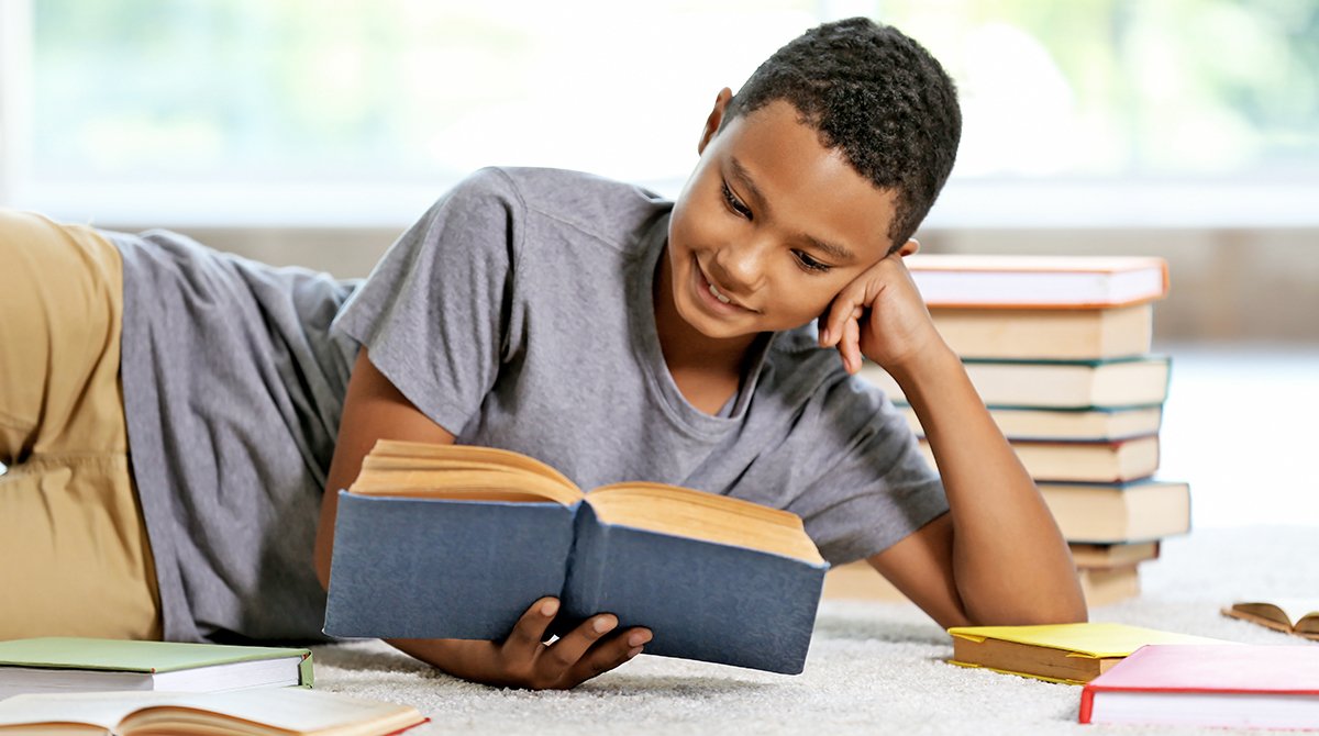 Photo of a young boy reading a book on the floor, surrounded by several other books.