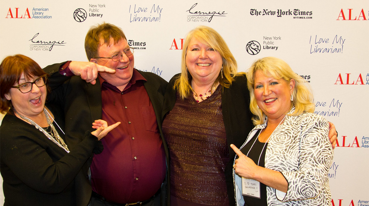 Photo of four happy people. Three of them are pointing to the woman in the middle. Logos in the background for the American Library Association, New York Public Library, The New York Times, Carnegie Corporation of New York, and the I Love My Librarian Award.