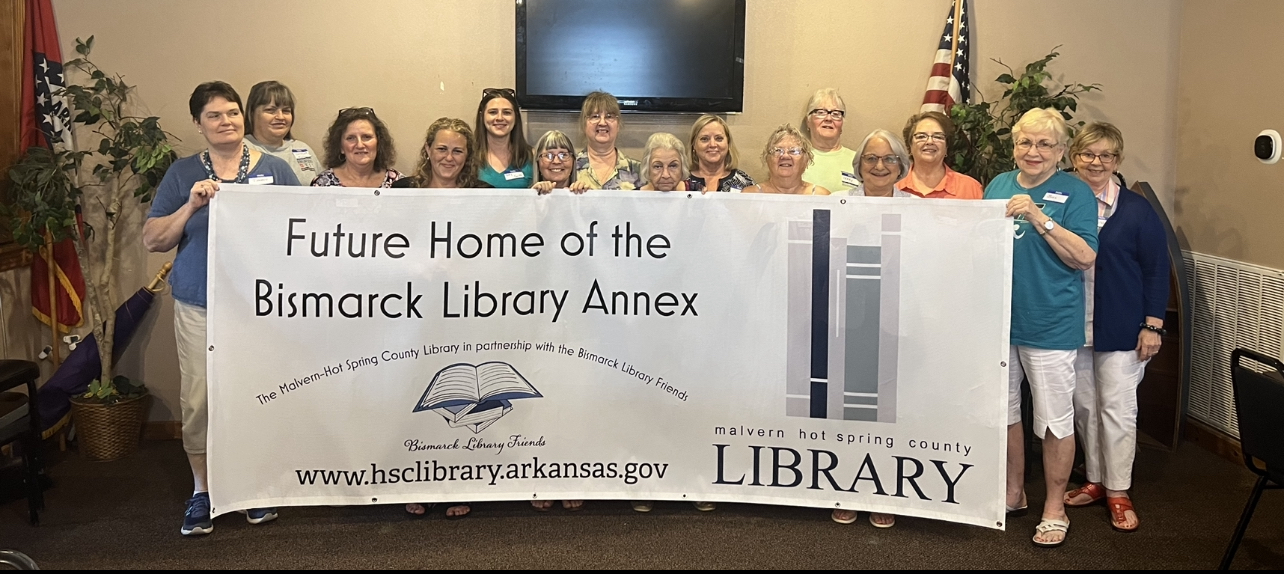 Photo of several people holding up a large banner that reads "Future Home of the Bismarck Library Annex. The Malvern-Hot Spring County Library in partnership with the Bismarck Library Friends. www.hsclibrary.arkasnas.gov."