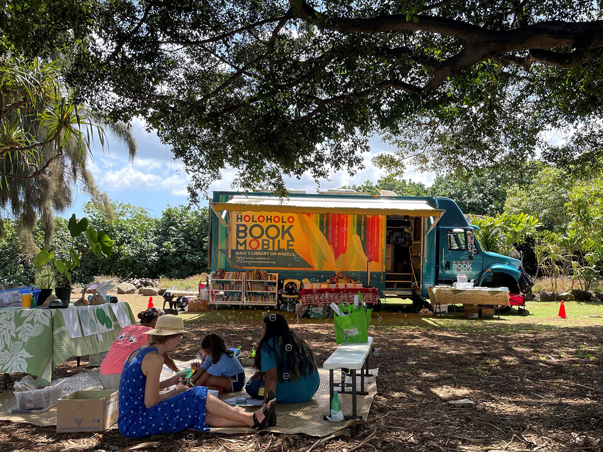 The Holoholo Bookmobile sits in a park as a family sits on the ground nearby.