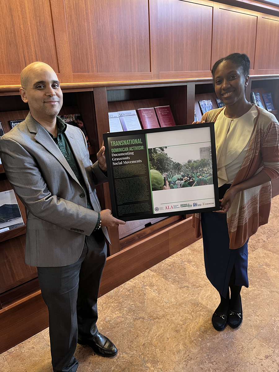 Two people holding up a framed poster from the exhibition "Transnational Dominican Activism: documenting Grassroots Social Movements"