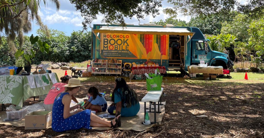 Photo of the Holoholo Bookmobile in a park and a family sitting on a blanket on the ground