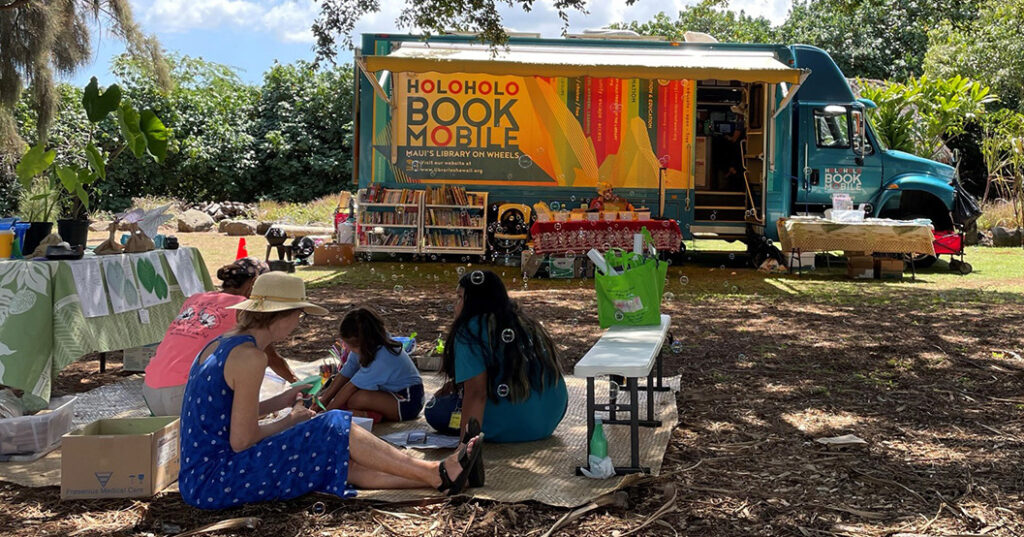 Photo of the Holoholo Bookmobile in a park and a family sitting on a blanket on the ground