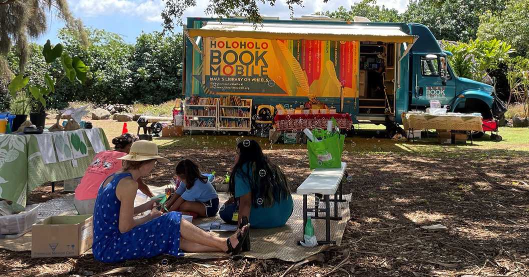 Photo of the Holoholo Bookmobile in a park and a family sitting on a blanket on the ground