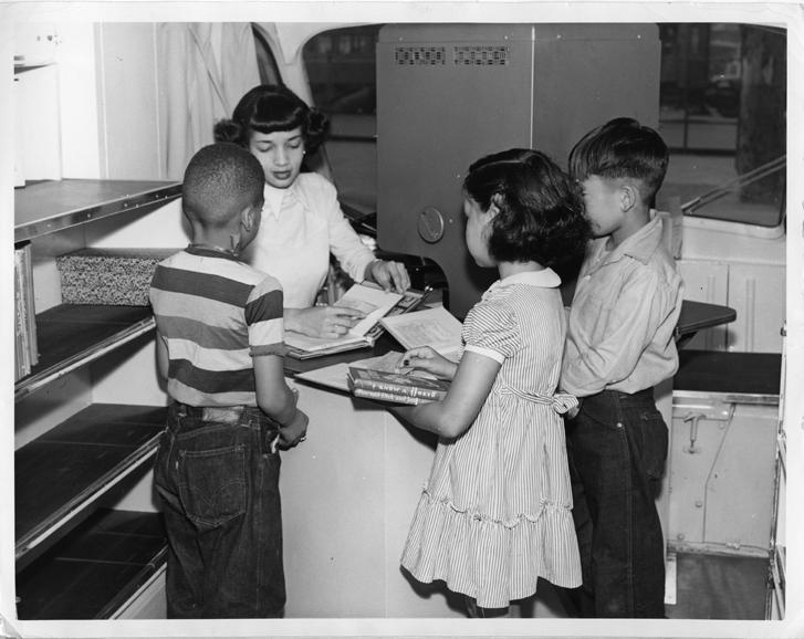 Original caption: "LOS ANGELES, October, 1949 – Elementary school pupils William Bias, Elizabeth Coggins and Jerry Sekido think it’s pretty interesting to watch Nellie Greene charge out their library books with the new Recordak charger on the Children’s Traveling Branch, operated by the Los Angeles Public Library.”