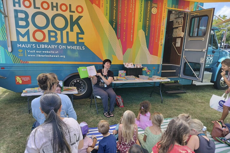 A librarian reading to a group of children at a park in front of the Holoholo bookmobile.