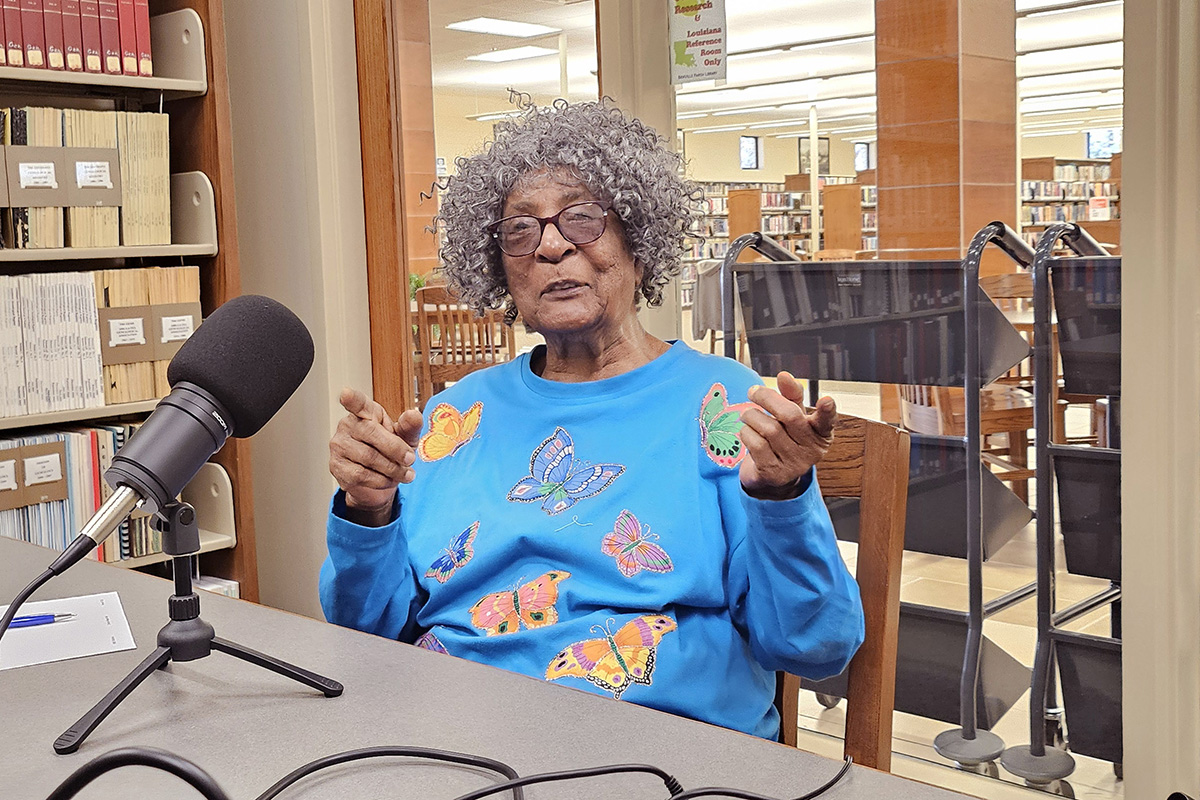 An elderly Black woman speaking into a microphone for an interview at a library