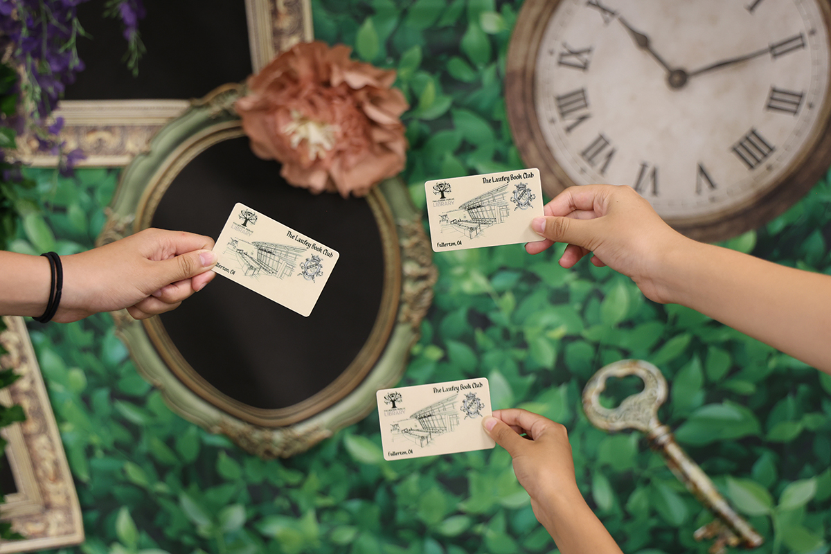 Photo of three arms holding out Laufey Book Club library cards in front of a backdrop with clocks and picture frames.