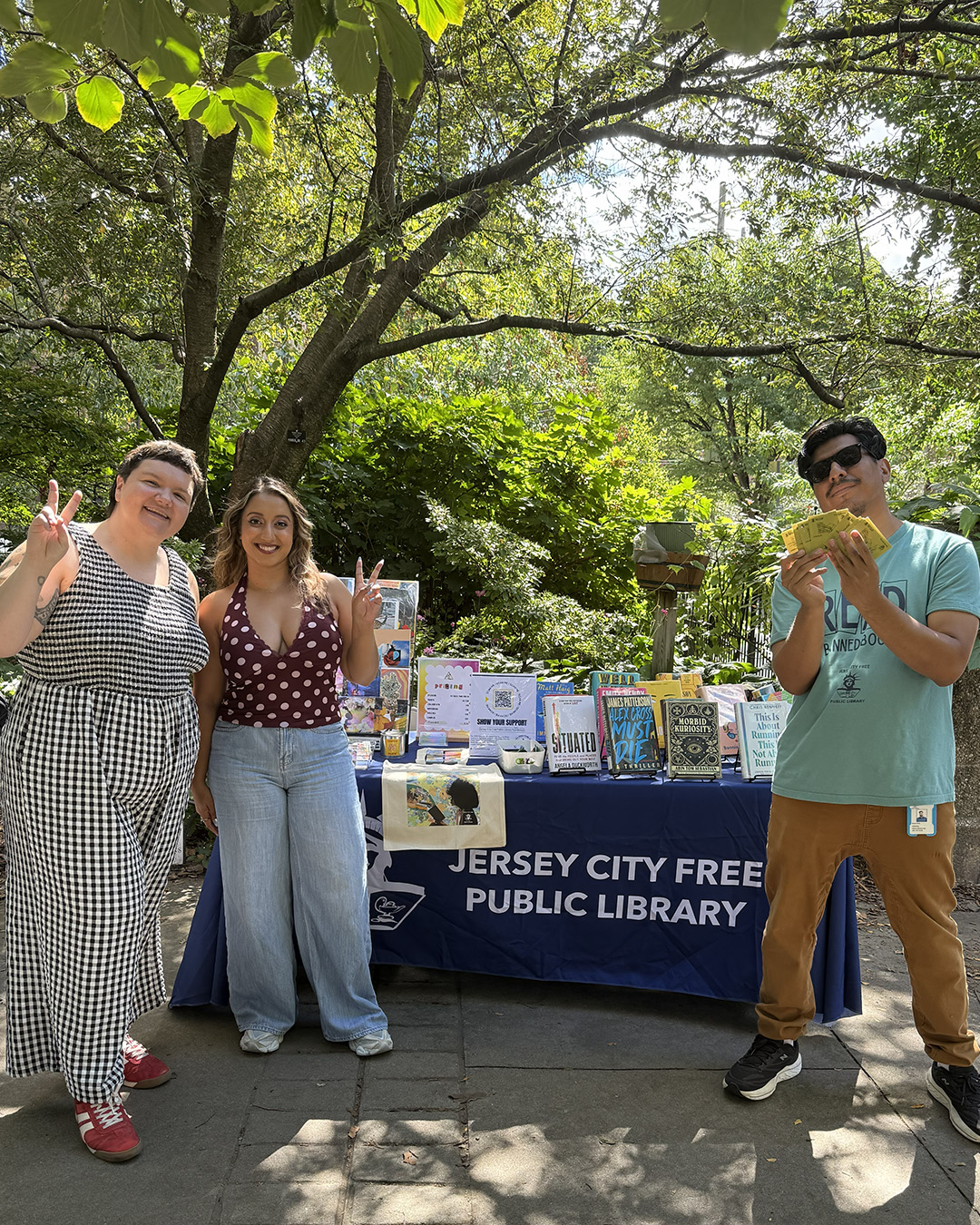 Photo of three people posing for a photo in front of a table for the Jersey City Free Public Library in the park/