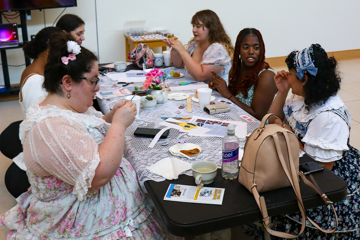 Photo of six young adult girls dressed in tea party dresses sitting at a crafting table in the library